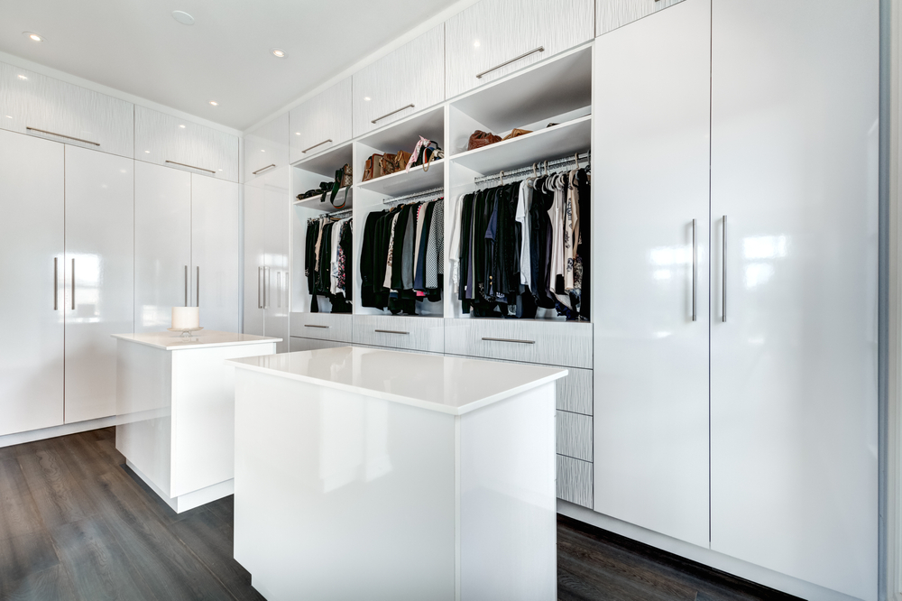 A walk in closet with multiple wardrobes and clothes hanging in the centre. The wardrobes are white with handles and gloss finish. 
